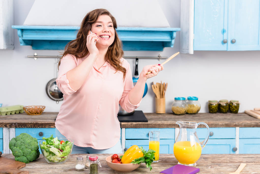 mujer joven con sobrepeso con cuchara de madera en la mano hablando en el teléfono inteligente mientras está de pie en la mesa con verduras frescas en la cocina en casa
 - Foto, imagen