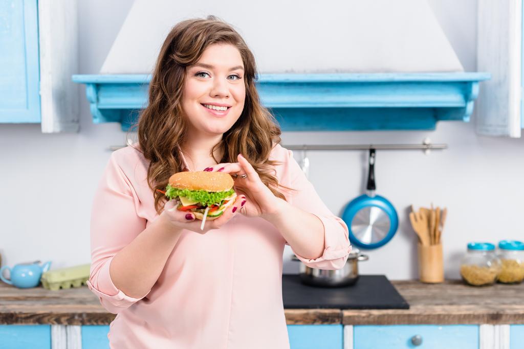 портрет молодої надмірної ваги жінки з бургер в руках на кухні вдома
 - Фото, зображення