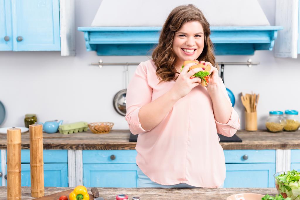 Porträt einer übergewichtigen lächelnden Frau mit Burger in der Hand in der heimischen Küche - Foto, Bild