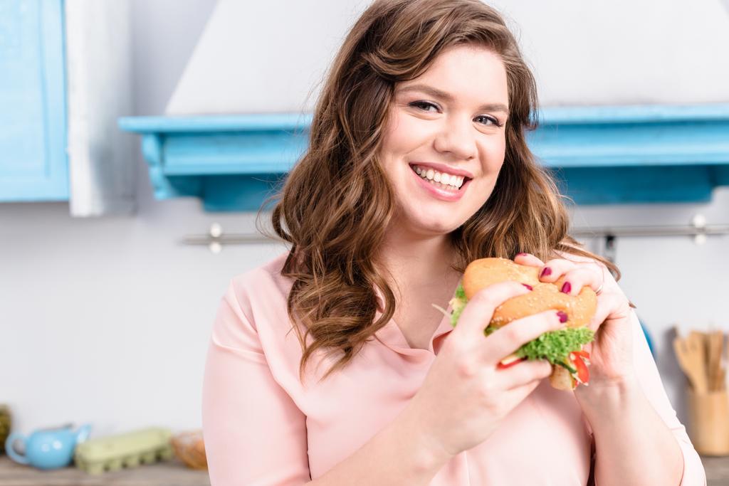 Porträt einer übergewichtigen lächelnden Frau mit Burger in der Hand in der heimischen Küche - Foto, Bild
