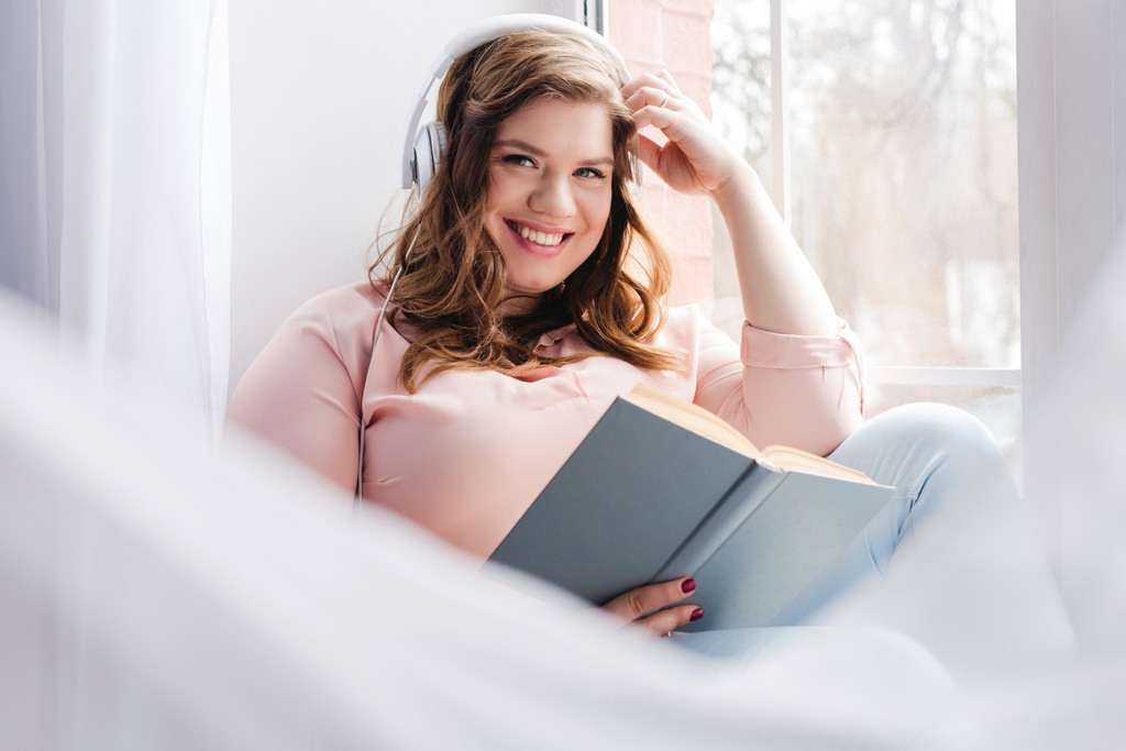usmívající se žena v sluchátka s knihou sedět na parapetu doma - Fotografie, Obrázek