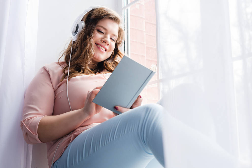 mujer joven en auriculares libro de lectura mientras está sentado en el alféizar de la ventana en casa
 - Foto, imagen