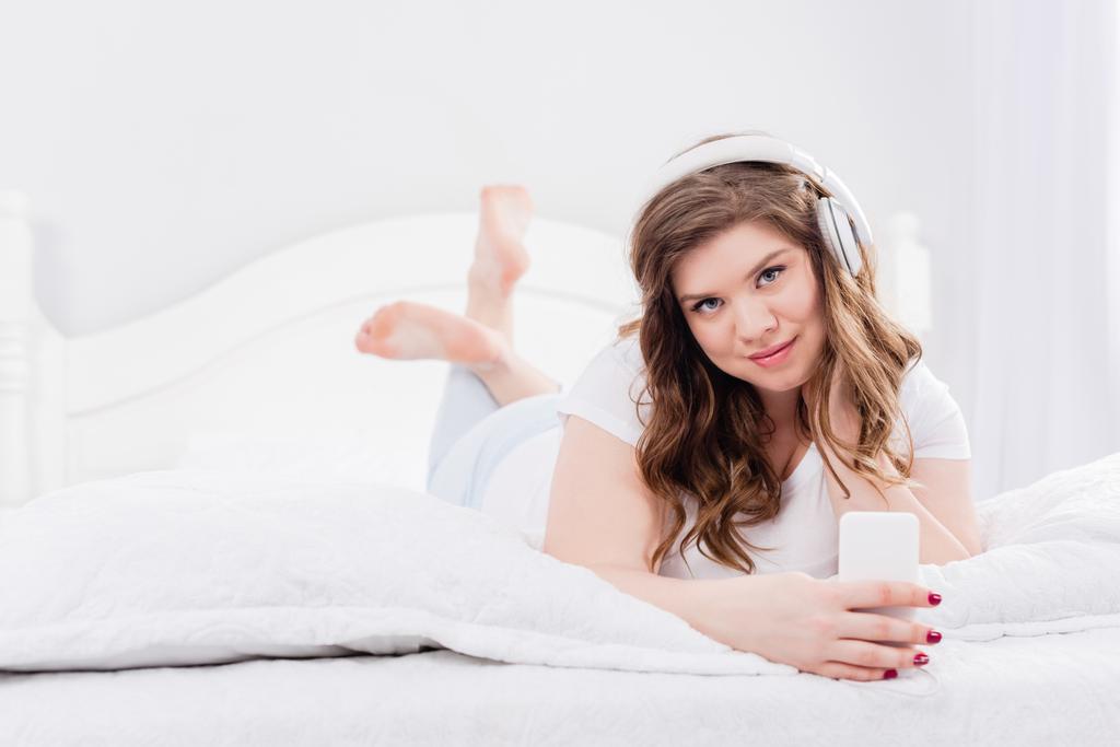 portrait of young woman in pajama and headphones with smartphone lying on bed at home - Photo, Image