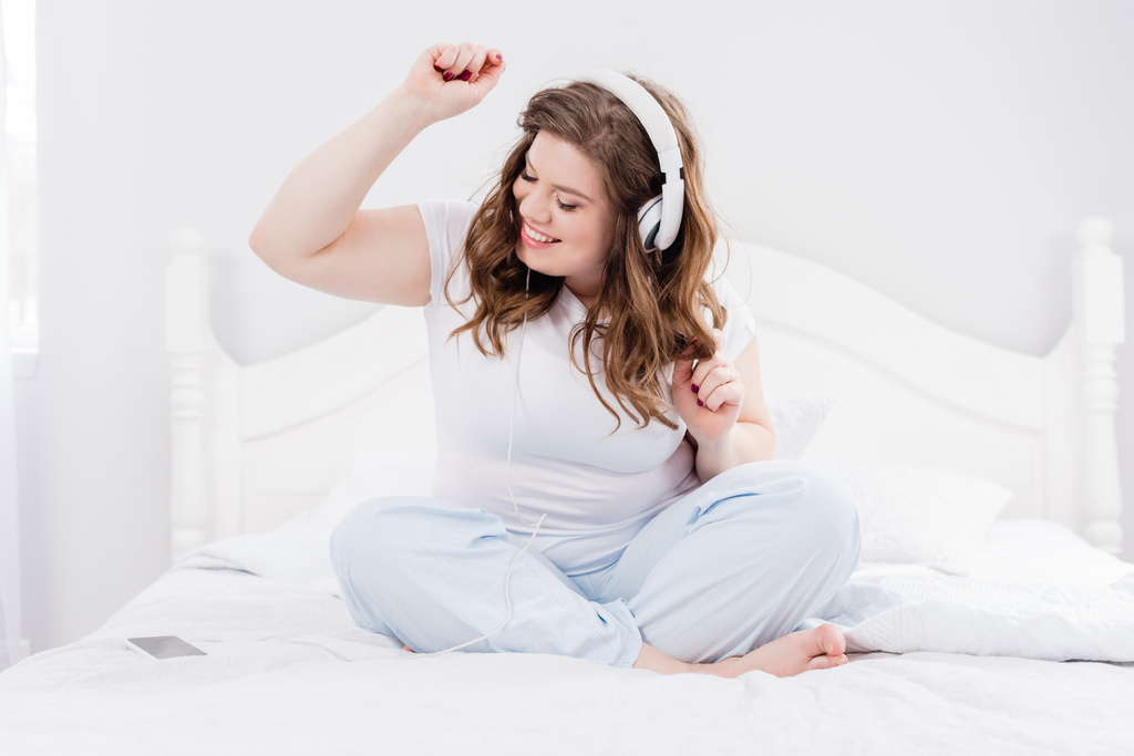 Lachende vrouw in pyjama luisteren muziek in hoofdtelefoons op bed thuis - Foto, afbeelding