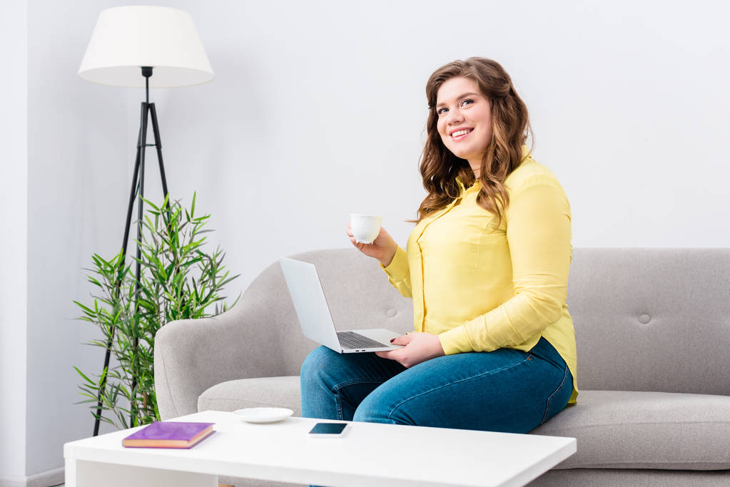 junge lächelnde Frau mit Tasse Kaffee und Laptop auf dem heimischen Sofa sitzend - Foto, Bild