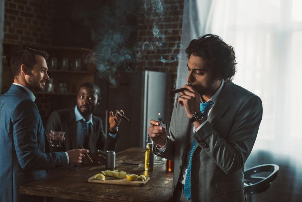 Красивый молодой бизнесмен курит сигары, а друзья пьют виски позади
  - Фото, изображение