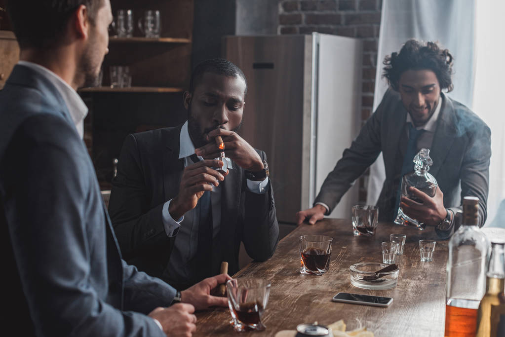 três homens multiétnicos de ternos fumando charutos e bebendo álcool juntos
 - Foto, Imagem
