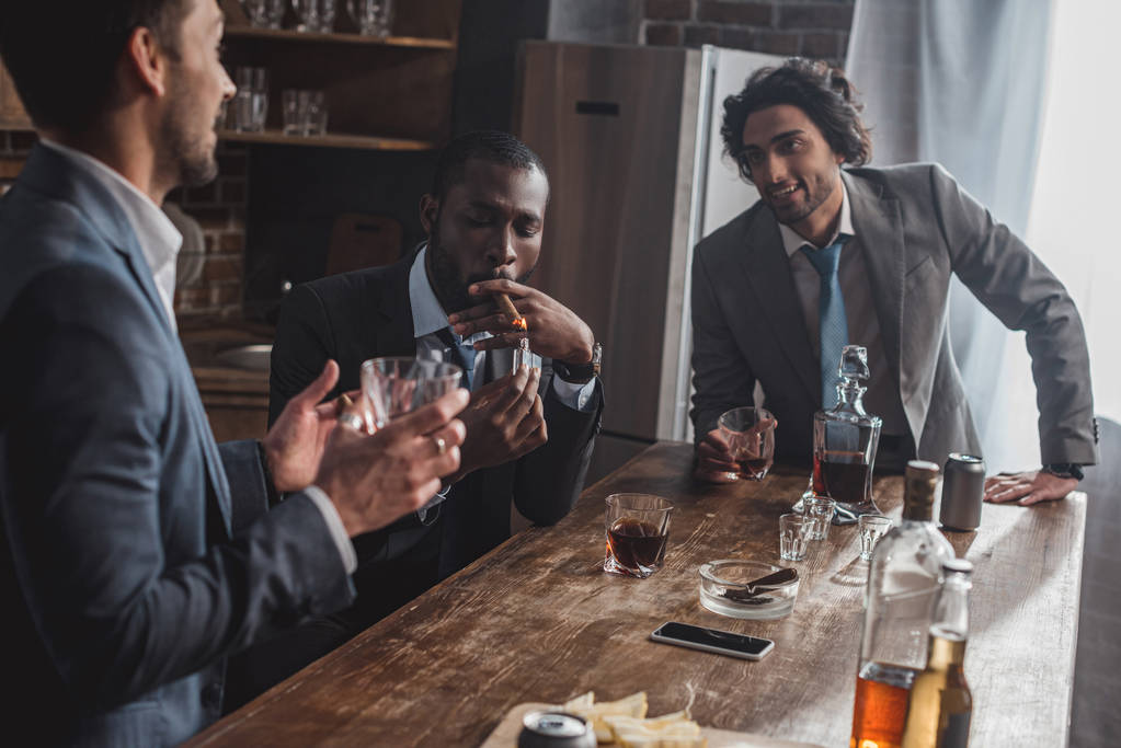 tiro recortado de homens de negócios multiétnicos sorridentes falando enquanto fumava charutos e bebia uísque
 - Foto, Imagem