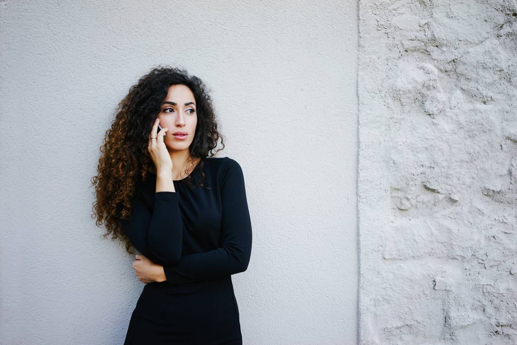 young beautiful businesswoman with long curly hair wearing a black dress and talking on phone against white brick wall background  - Photo, Image