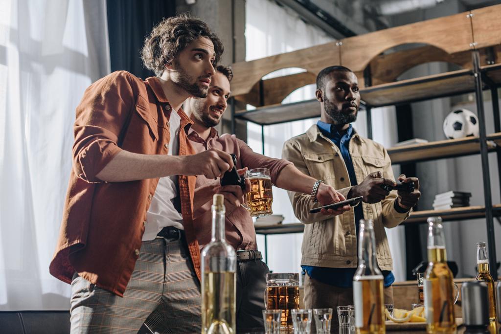 niski kąt widzenia wieloetnicznym mężczyzn, grając z joysticków i picie piwa razem - Zdjęcie, obraz