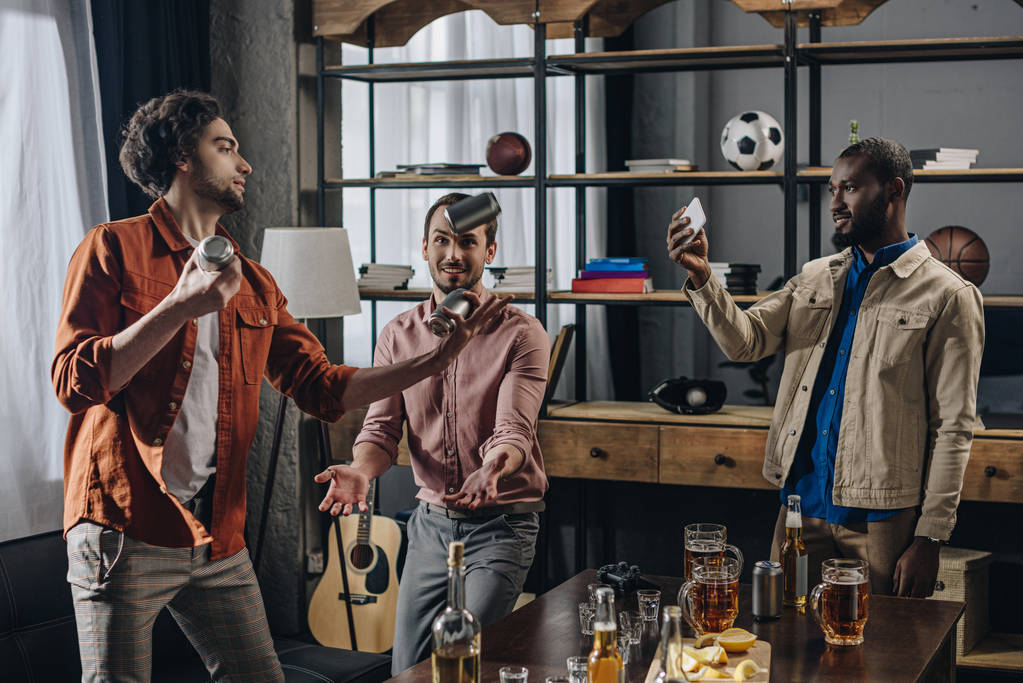 uomo afroamericano utilizzando smartphone e guardando gli amici giocoleria con lattine di birra
 - Foto, immagini