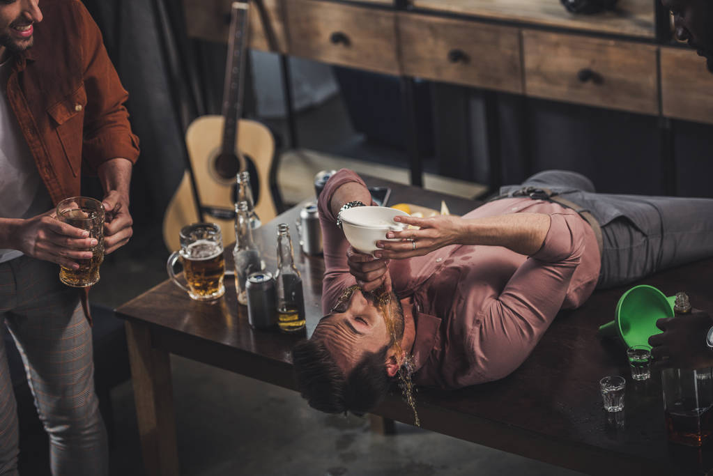 jonge man liggend op tafel en het drinken van bier uit de trechter terwijl vrienden kijken naar hem - Foto, afbeelding