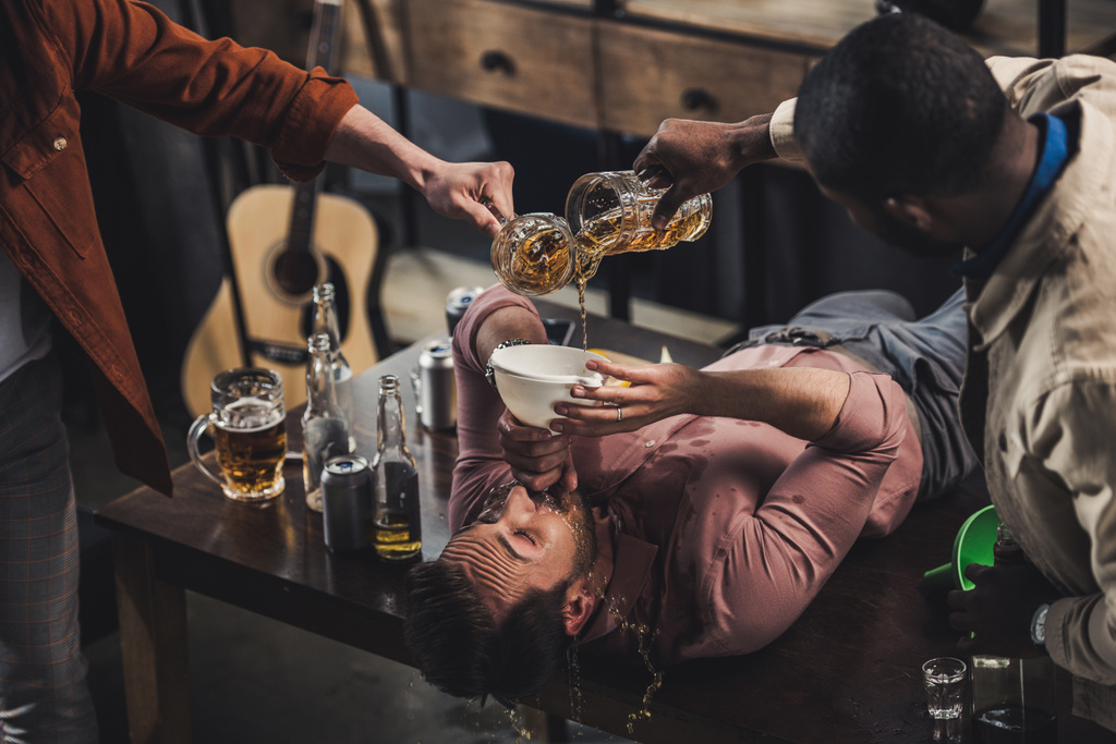 Schnappschuss von Freunden, die Bier in Trichter schütten und Mann trinkt, während er auf Tisch liegt - Foto, Bild