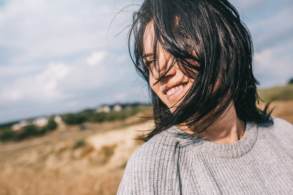 Retrato recortado horizontal de la hermosa mujer sonriente que usa suéter que es juguetona con el pelo ventoso y sin preocupaciones posando sobre el cielo de la luz del sol y el fondo de la naturaleza. Viajes, personas y estilo de vida
. - Foto, Imagen