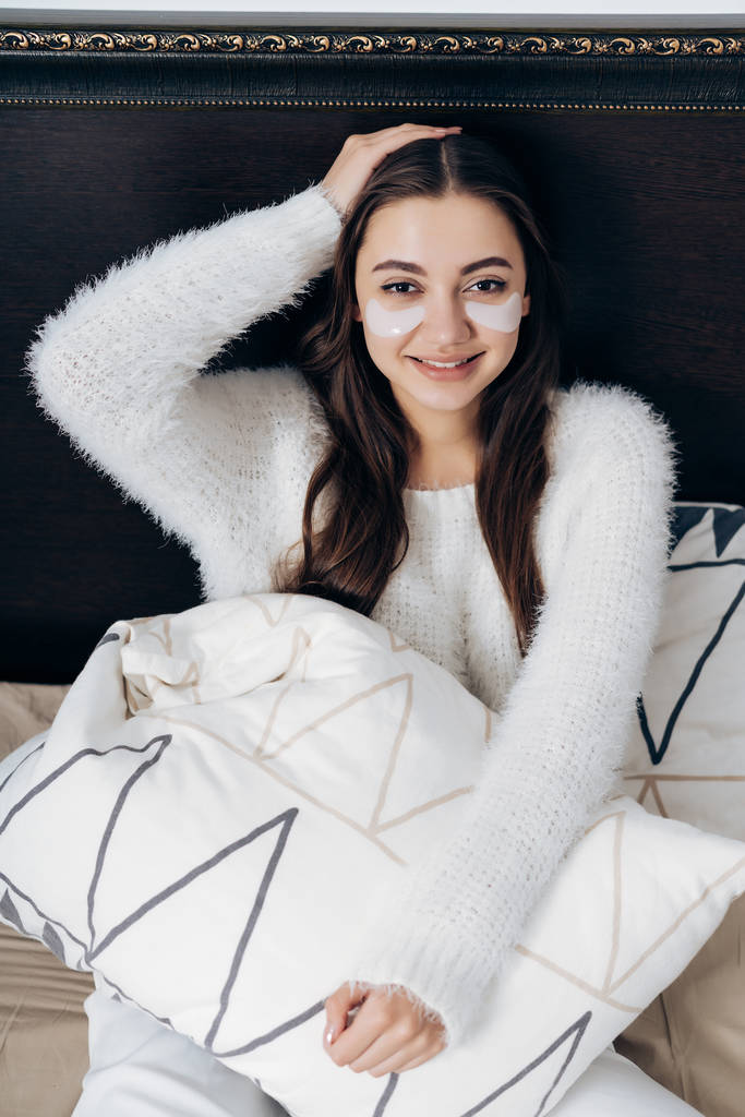 bela menina de cabelos longos em pijama branco senta-se na cama, no início da manhã, sob os olhos de manchas brancas, sorrindo
 - Foto, Imagem