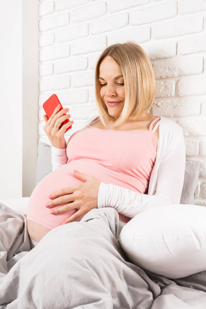 Gelukkig lachende zwangere vrouw ligt in bed en kijkt naar mobiele telefoon - Foto, afbeelding