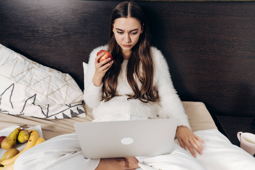 seriózní dlouhé vlasy dívka v pyžamu leží v posteli, vypadá zajímavé epizody na notebooku a žere jablko - Fotografie, Obrázek