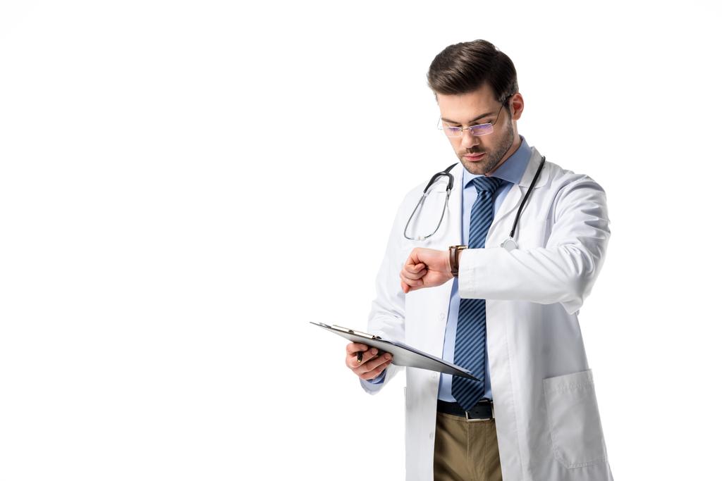 Arzt mit Klemmbrett trägt weißen Mantel mit Stethoskop und überprüft seine Uhr isoliert auf weiß - Foto, Bild