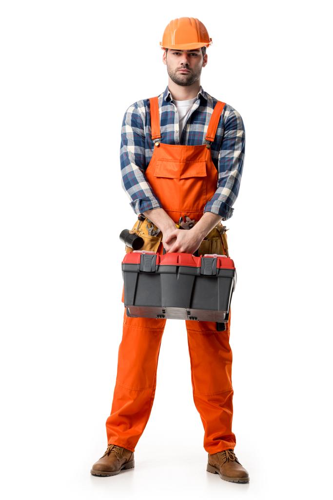 Handyman confiante em laranja geral e chapéu duro segurando caixa de ferramentas isolado no branco
 - Foto, Imagem