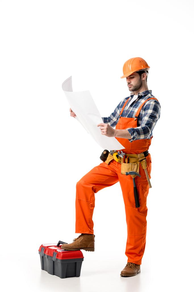 Bauarbeiter in orangefarbenem Overall und Helm blickt auf Blaupause, während er sich auf Werkzeugkiste stützt - Foto, Bild