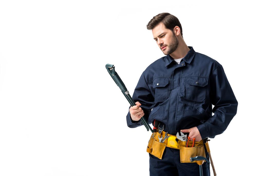 Ingeniero sanitario barbudo que usa uniforme con cinturón de herramientas y mira la llave inglesa aislada en blanco
 - Foto, Imagen