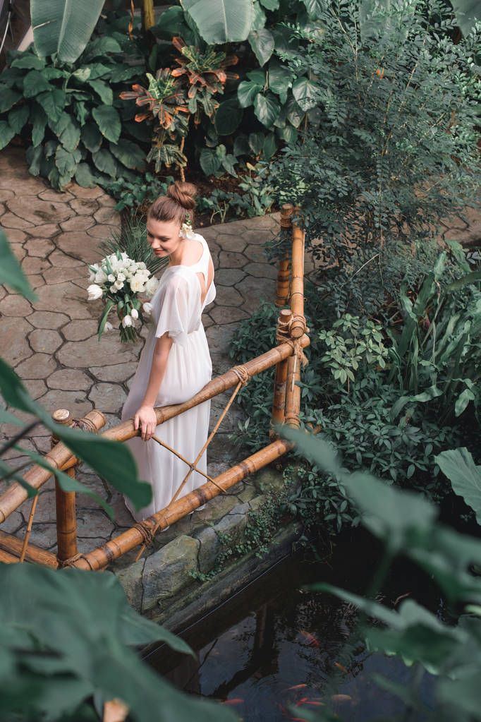 Blick von oben auf attraktive Braut posiert in weißem Kleid mit Brautstrauß im tropischen Garten - Foto, Bild