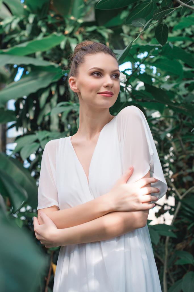 atrakcyjna Panna Młoda w białej sukni z skrzyżowanymi rękami pozowanie w tropikalnym ogrodzie - Zdjęcie, obraz