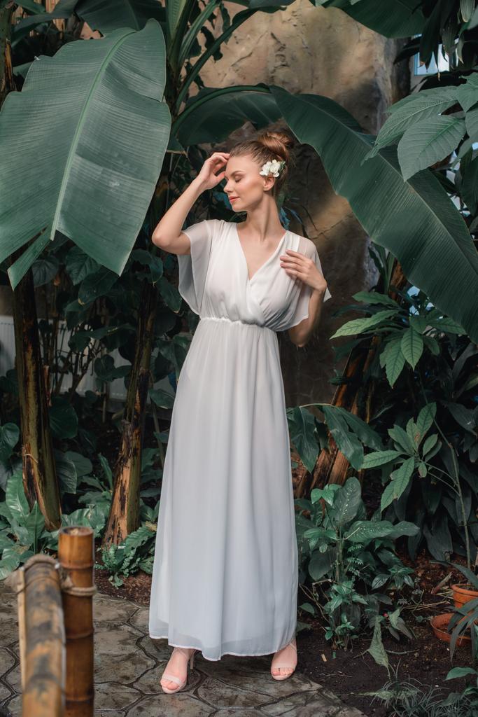 bonita mujer joven en vestido blanco con flor en el pelo posando en jardín tropical
 - Foto, Imagen