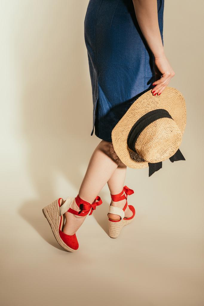 imagem cortada de mulher elegante em sandálias plataforma vermelha segurando chapéu de palha no fundo bege
 - Foto, Imagem