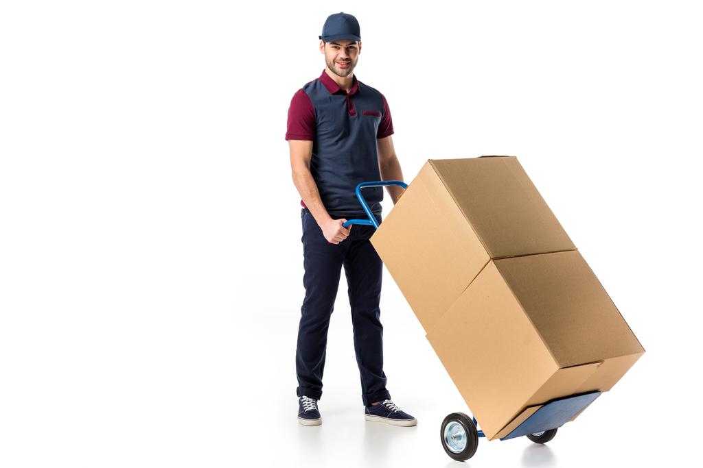 glimlachend levering man in uniform duwen hand vrachtwagen met kartonnen dozen geïsoleerd op wit - Foto, afbeelding