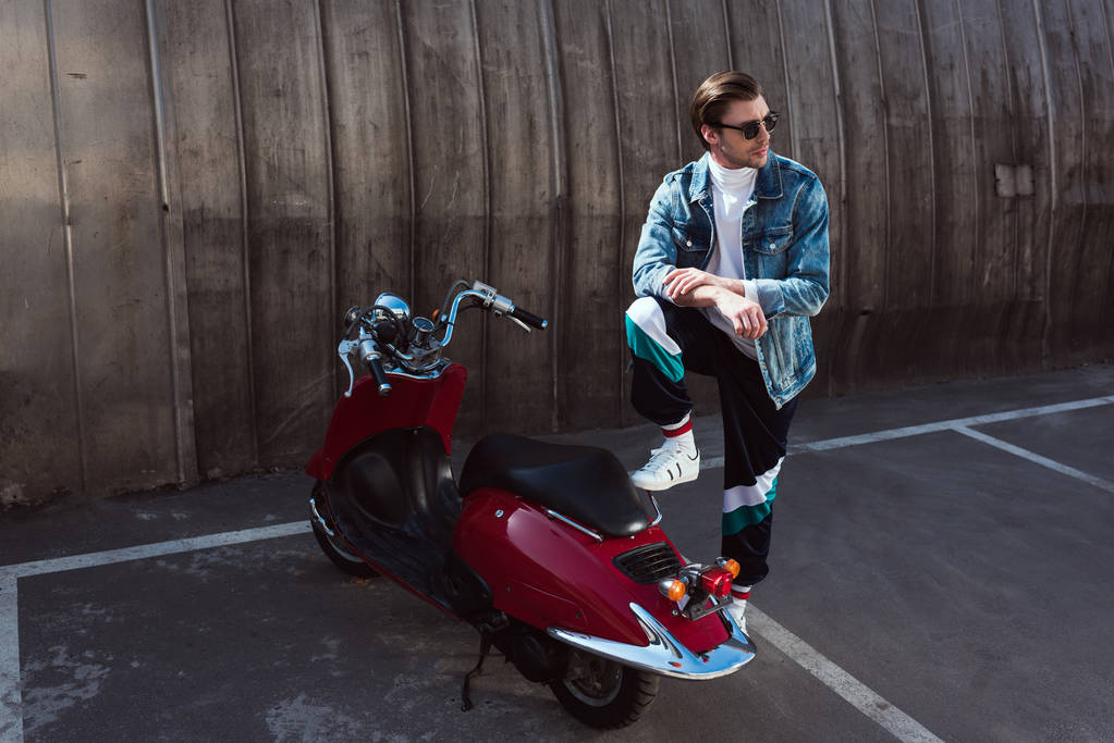 knappe jongeman in stijlvolle denim jasje met vintage scooter op parking - Foto, afbeelding