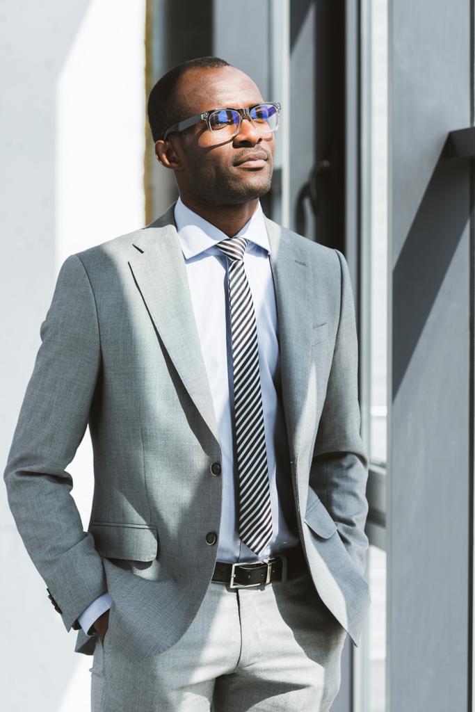 уверенный в себе молодой африканский американский бизнесмен в очках стоит с руками в карманах и смотрит в сторону
 - Фото, изображение