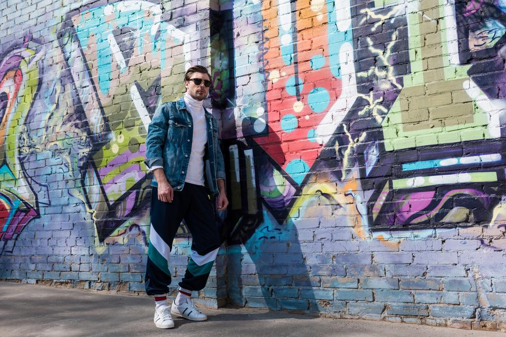 elegante giovane uomo in giacca di denim vintage e pantaloni da pista appoggiati su muro di mattoni con graffiti
 - Foto, immagini