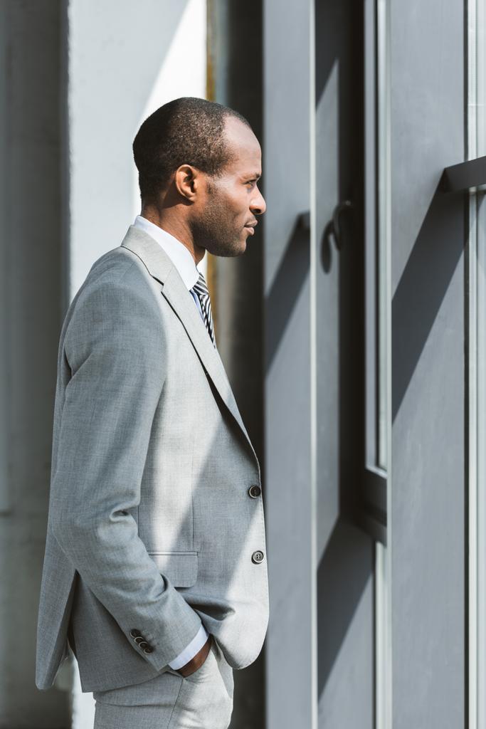 Zijaanzicht van lachende jonge Afro-Amerikaanse zakenman permanent met hand in de zak en kijken naar venster - Foto, afbeelding