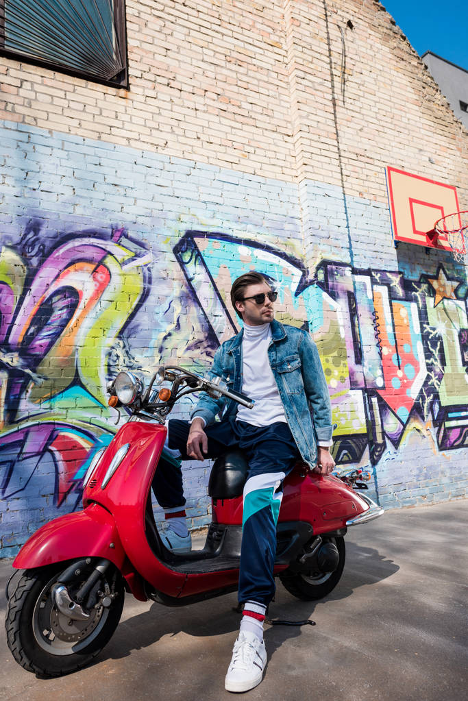 joven guapo en ropa elegante en scooter rojo vintage en frente de la pared de ladrillo con graffiti
 - Foto, imagen