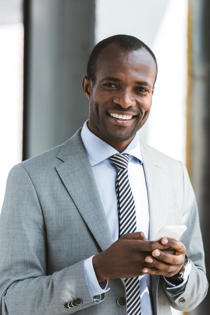 Portret van knappe jonge Afro-Amerikaanse zakenman met behulp van smartphone en lachend op camera - Foto, afbeelding
