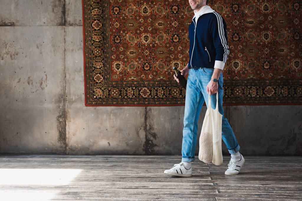 cropped shot of man in old school clothes with string bag and bottle of beer in front of rug hanging on wall - Photo, Image