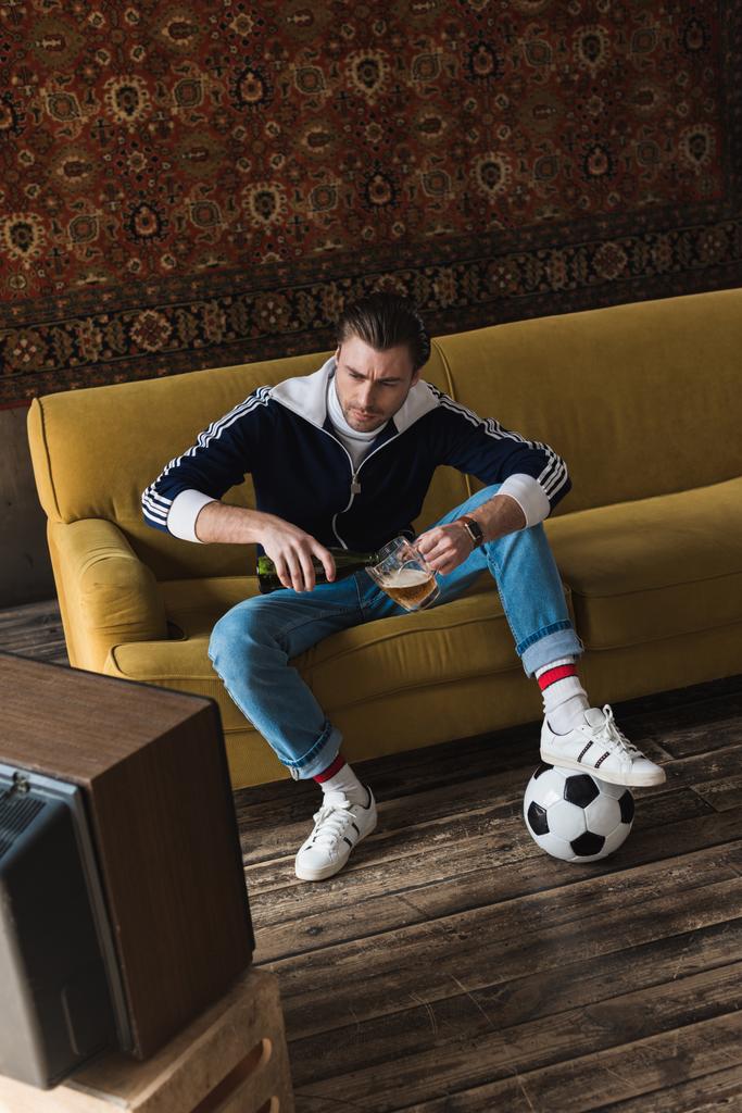 serious young man in vintage clothes with ball watching soccer on old tv and pouring beer into mug - Photo, Image