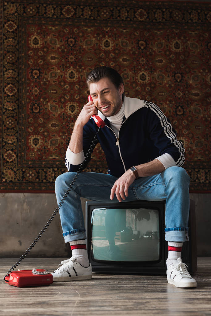 vintage giyim eylem ayarlayıp önünde duvara asılı halı telefonla konuşurken retro TV oturan genç adam gülümseyen - Fotoğraf, Görsel
