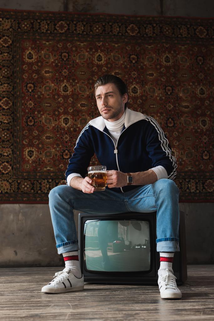 stylischer junger Mann in Vintage-Klamotten mit einem Becher Bier sitzt auf einem Retro-Fernseher vor Teppich an der Wand hängen - Foto, Bild