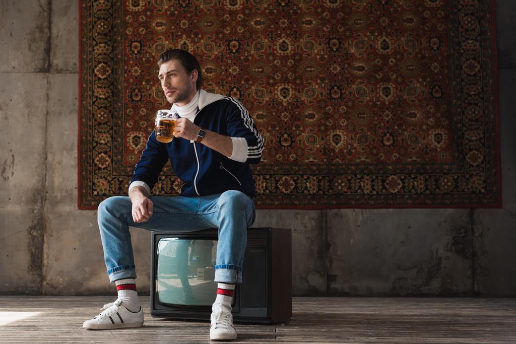zadumany młodzieniec w ubrania vintage z kuflem piwa, siedząc na retro tv ustawiony przed dywan, wiszące na ścianie - Zdjęcie, obraz