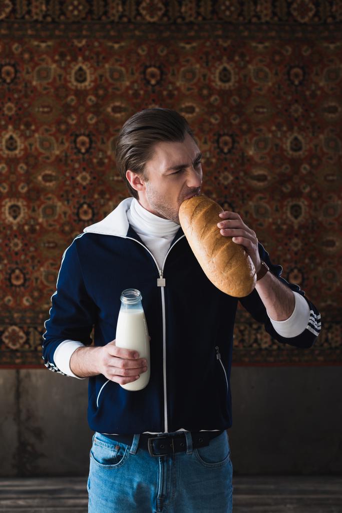 νεαρός άνδρας στο vintage ρούχα με μπουκάλι γάλα δάγκωμα καρβέλι ψωμί μπροστά από την κουβέρτα που κρέμονται στον τοίχο - Φωτογραφία, εικόνα
