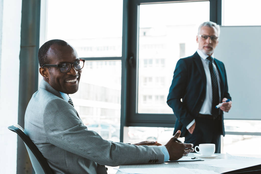przystojny biznesmen Afroamerykanów w okularach, uśmiechając się aparatu podczas pracy z mężczyzna kolegi w biurze - Zdjęcie, obraz