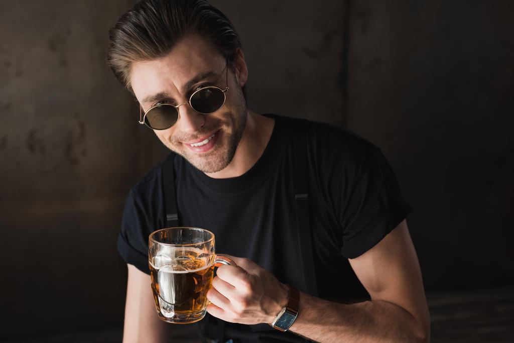 joven sonriente en camiseta negra y gafas de sol sosteniendo taza de cerveza y mirando a la cámara
 - Foto, imagen