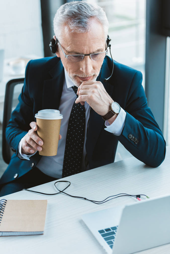 homme d'affaires senior dans les lunettes et le casque à l'aide d'un ordinateur portable et tenant tasse en papier
 - Photo, image
