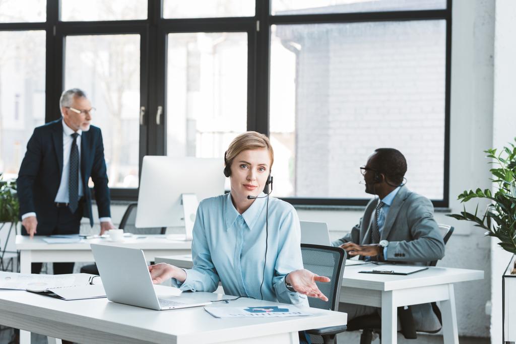 Geschäftsfrau im Headset mit Laptop und Blick in die Kamera, während männliche Kollegen im Büro hinten arbeiten - Foto, Bild