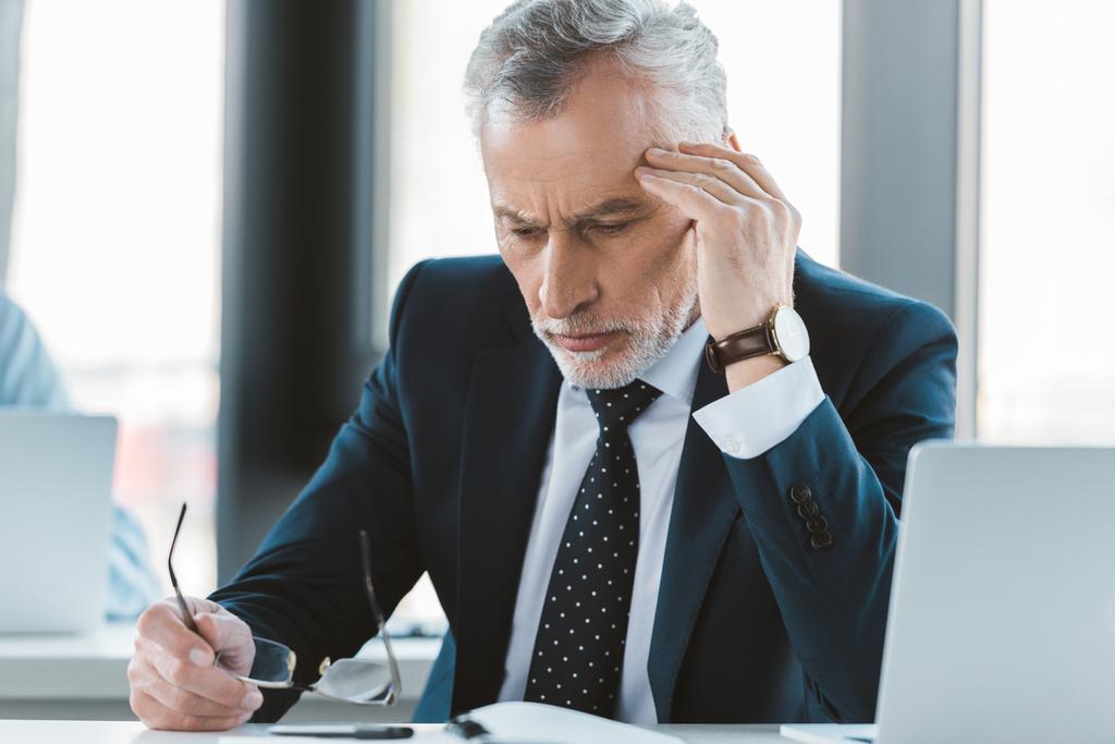 moe senior zakenman met hoofdpijn bril houden en kijken neer op de werkplek - Foto, afbeelding