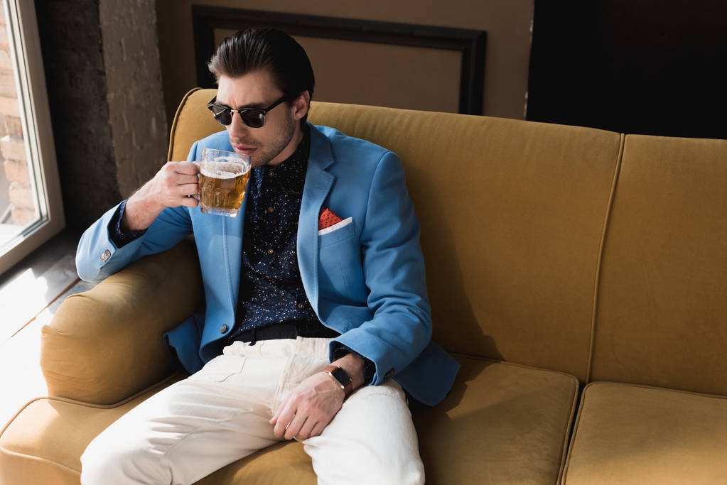 viehättävä nuori mies tyylikkäässä puvussa istuu sohvalla ja juo olutta
 - Valokuva, kuva