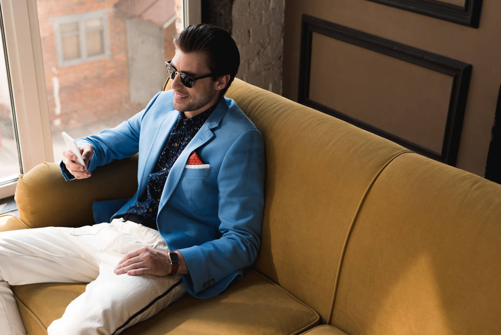 Blick aus der Vogelperspektive auf einen lächelnden jungen Mann im eleganten Anzug mit Smartphone, während er auf der Couch sitzt - Foto, Bild