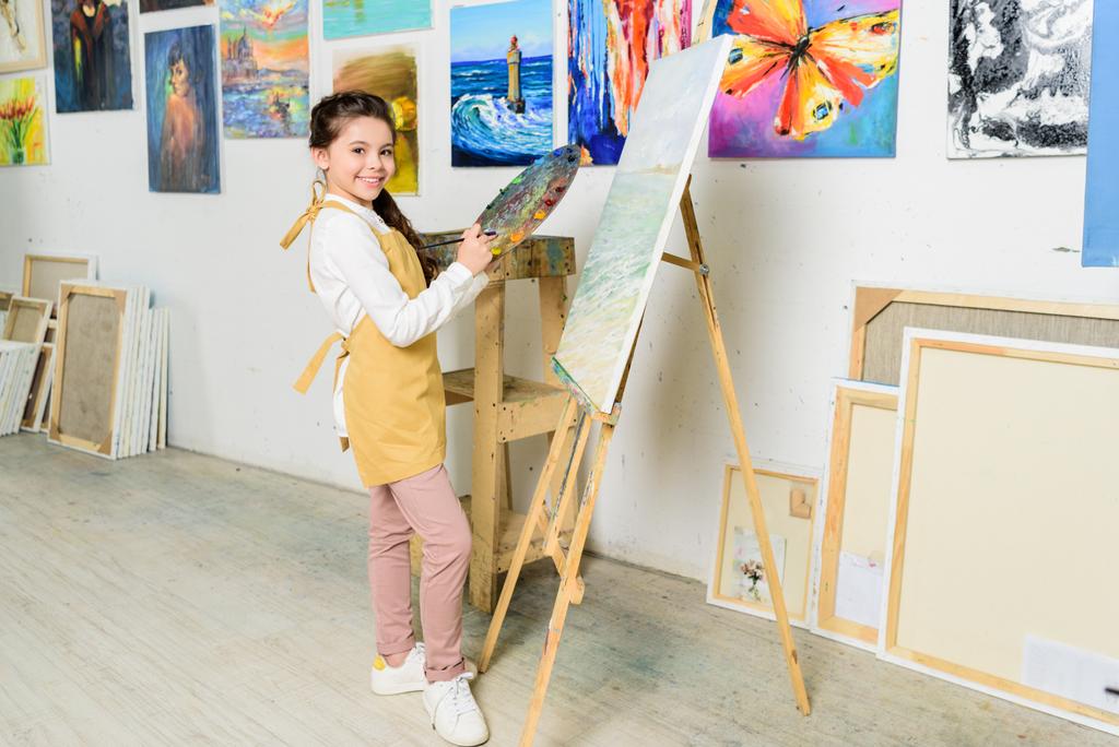 niño sonriente de pie cerca de lienzo en el taller de la escuela de arte y mirando a la cámara
 - Foto, Imagen